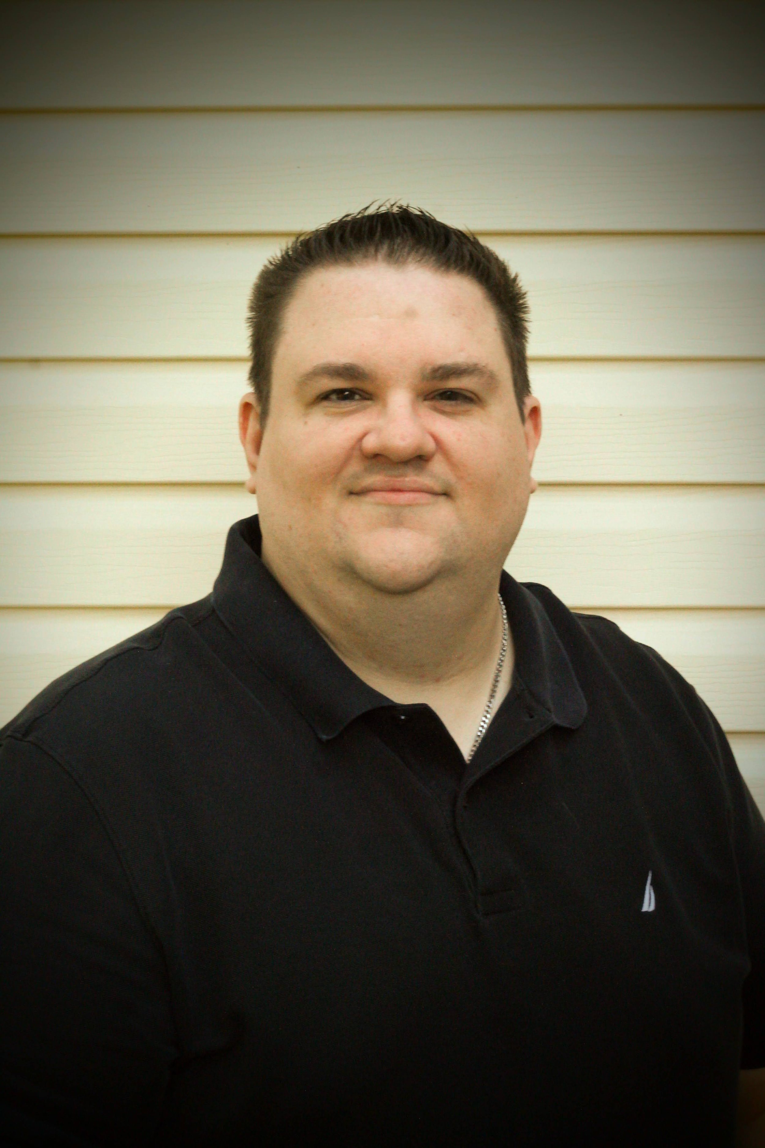Photograph of a man with short dark hair wearing a black polo shirt, standing outdoors against a light beige horizontal wooden siding background.