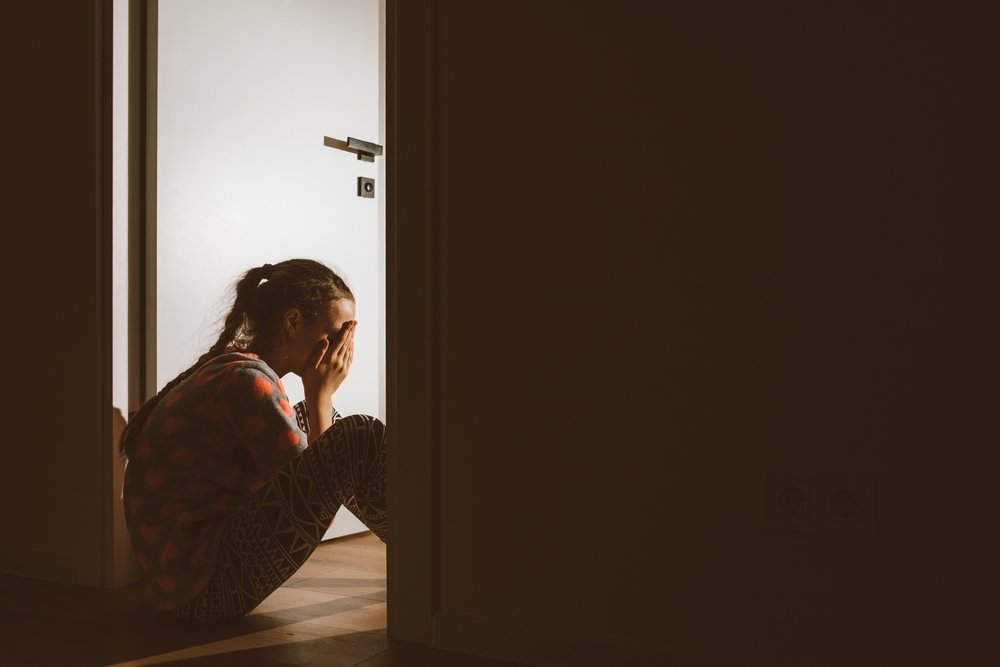 A young girl sits on the floor in a dimly lit room, holding her head in her hands near a bright doorway, appearing distressed or upset.